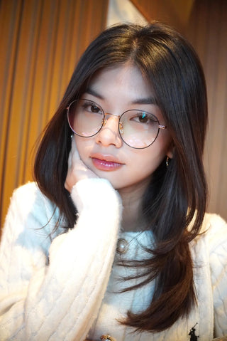 A young woman with long dark hair wears EyeCandys Luna Round eyeglasses as she rests her chin on her hand, gazing neutrally at the camera in a white sweater. Brown vertical blinds form the background.