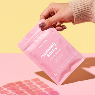 A hand with painted nails holds a pink pouch labeled "Insanely Sticky Tabs for Press-On Nails (390 pc)" by EyeCandys against a yellow background. Below, MMA-free adhesive sheets are shown on a pink surface.