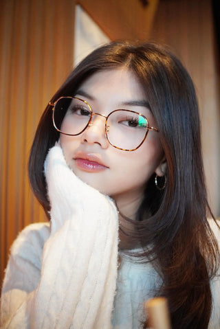 A young woman with long dark hair and round glasses rests her chin on her hand, wearing a white sweater. She showcases EyeCandys Farrow Round glasses in a softly lit indoor setting, looking at the camera with a relaxed expression.