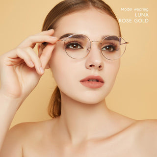 A woman with light skin and brown hair wears EyeCandys Luna Round vintage-style titanium glasses in rose gold, holding the frames. She has natural makeup and a neutral background. Text reads: "Model wearing LUNA ROSE GOLD.