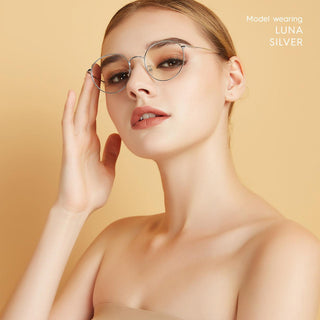A woman with fair skin and slicked-back hair poses against a beige background, wearing EyeCandys Luna Round titanium glasses with round frames and minimal makeup. Text in the corner reads “Model wearing LUNA ROUND.”.
