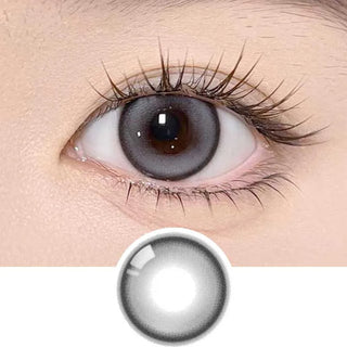 A close-up of an eye wearing Olola Meow Natural Coon Grey contacts from Olola, which enlarge the iris and feature long lashes. A single grey Olola lens is displayed separately below the eye.