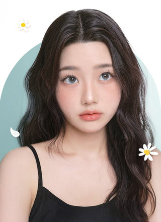 A young woman with long wavy dark hair and blue-green eyes, enhanced by i-Girl One Pick Becca Matcha Blue contact lenses, looks upward. She wears a black tank top and soft makeup, surrounded by cartoon daisies on a light blue background.