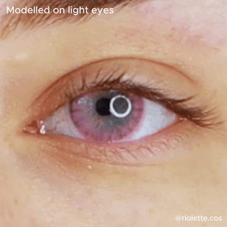 A close-up of a light-colored eye wearing EyeCandys Shade Pink contact lens. The text reads "Modeled on light eyes." Credit: @riolette.cos in the lower right. The pupil reflects a ring light. Ideal for natural-looking pink contacts.