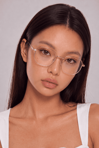 A young woman with long dark hair and glasses looks at the camera with a neutral expression, wearing EyeCandys Stunner Square metal frame glasses and a white top against a light gray background.