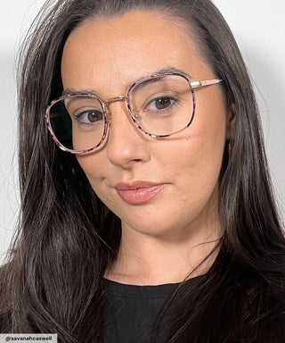 A woman with long dark hair and clear skin models EyeCandys Stunner Square eyeglasses in metal frames, wearing a black top against a white background. She poses with a neutral expression, highlighting these chic fashion glasses.