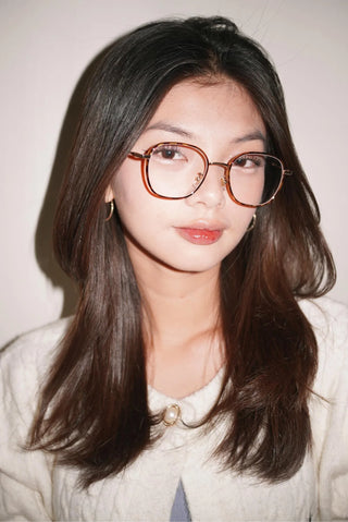 A young woman with long, straight dark hair wears EyeCandys’ Farrow Round vintage glasses and a cream textured jacket with a pearl brooch, gazing calmly at the camera against a simple light background.