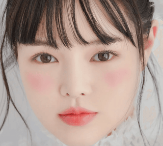 Close-up of a young woman with smooth, fair skin, rosy cheeks, and soft pink lips. She has straight black hair with bangs and is wearing a white lace top. Her expression is calm and serene.