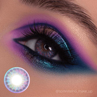 A close-up of an eye with vibrant purple, blue, and teal eyeshadow, a dramatic black eyeliner, bold lashes, and a colorful contact lens with a sunburst pattern.