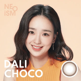 A woman with shoulder-length dark hair and a warm smile poses in front of a peach background, wearing a sleeveless orange top. Beside her is an image of NEO Vision Neoism Dali Choco 1-Day (50pk) daily disposable contact lenses.