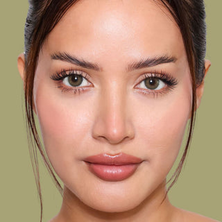 A woman with smooth, glowing skin and natural makeup, featuring full lips, defined eyebrows, and long eyelashes, looks directly at the camera against a plain, muted olive background.