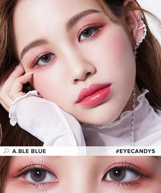 A close-up of a woman with flawless skin, pink eyeshadow, and bold pink lips wearing Olola Able Blue color contact lenses by Olola for dark eyes. Her hand touches her face as a chain earring dangles. Below is a zoom-in of the vibrant blue lenses.