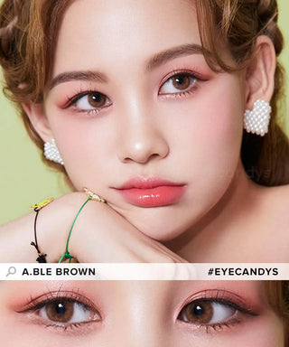 A woman with brown eyes, peachy makeup, and glossy pink lips poses with her chin on her arm, wearing pearl earrings and beaded bracelets. A close-up highlights her Olola Able Brown color contact lenses for dark eyes by Olola (#EYECANDYS).