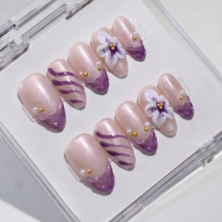 The EyeCandys Aloha Darling Press-On Nails set features glossy, pale pink nails with purple tips, accented by diagonal stripes, gold beads, and 3D white and purple flower details, all neatly arranged in a clear case.