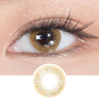 A close-up of an eye with long lashes and shimmery gold makeup wearing EyeCandys Attitude Glitter Brown color contact lenses. Below, a circular image highlights the natural design of these EyeCandys contacts.