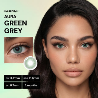 A woman with long dark hair wears EyeCandys Aura Green Grey contacts in close-up. These UV-blocking lenses create a jewel-toned eye effect. Specs: DIA 14.2mm, G.DIA 13.6mm, B.C. 8.7mm, usable for 3 months. "EyeCandys Aura Green Grey" appears onscreen.