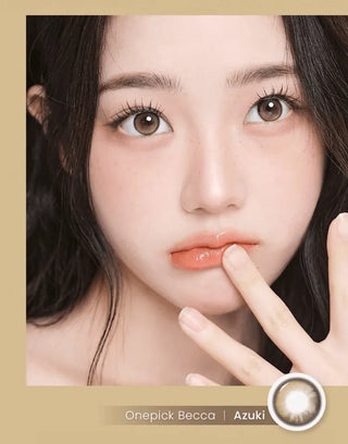 A woman with long dark hair and brown eyes gazes at the camera, fingers near her lips. She wears natural makeup, glossy peach lipstick, and i-Girl One Pick Becca Azuki Brown contact lenses by i-Girl shown at the bottom right.