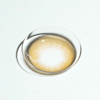 The EyeCandys Pink Label Barcelona Brown contact lens, featuring a light center and dark rim, is displayed on a white surface—ideal for anyone wanting natural-looking brown contacts.
