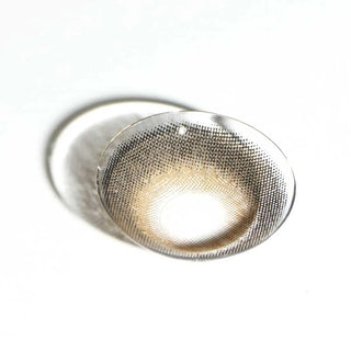 A single EyeCandys Pink Label Big Ring Brown lens with a brown and black dotted pattern rests on a white surface, casting a soft shadow—ideal for doll-like, eye-enlarging effects.