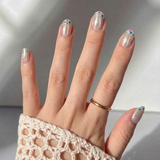 A hand wears a gold ring and a cream crochet sleeve. The Birthday Cake Press-On Nails by EyeCandys feature shimmery polish with playful, colorful flower designs near the tips for a fun, festive look.