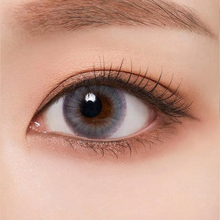 A close-up of a human eye with brown brows and long lashes wearing Olola Blow Midnight Grey by Olola, which gives dark eyes a striking blue-grey iris effect. The skin around the eye appears naturally smooth.