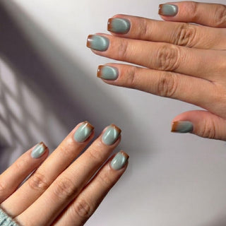 Two hands display EyeCandys Blue Hour Latte Press-On Nails—short, square-shaped and light sage green with a thin brown border—shown against a plain white background with soft shadows.