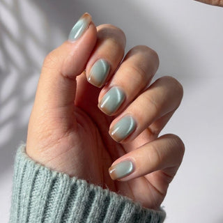 A hand with short, neatly manicured EyeCandys Blue Hour Latte Press-On Nails in a shimmery pale green is shown. The person wears a light green knitted sweater, with soft natural light and gentle shadows in the background.