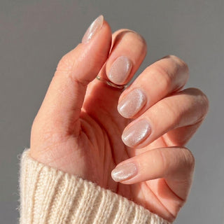 A hand wears EyeCandys Business Casual Press-On Nails in shimmering nude, held against a neutral background. The person is dressed in a cream-colored knit sweater.