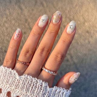 A hand wears EyeCandys Constellation Prize Press-On Nails, featuring pastel Y2K art with white stars, metallic stickers, sheer glitter, gold rings, and a white knit sleeve.