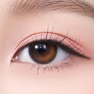 A close-up of a person's eye with natural makeup and long eyelashes, wearing Olola Daymood Brown myopia prescription contact lenses by Olola. The skin around the eye appears smooth and softly lit.