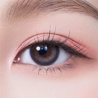 A close-up of a human eye with natural makeup and long eyelashes, wearing Olola Daymood Grey color contact lens by Olola for dark eyes. The eyebrow and smooth skin around the eye are visible.