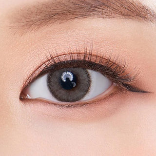 A close-up of a person's eye wearing Olola Dearsome Ash Grey contact lenses by Olola, featuring natural-looking makeup, long lashes, and clear details of the iris and surrounding skin—ideal for dark eyes.