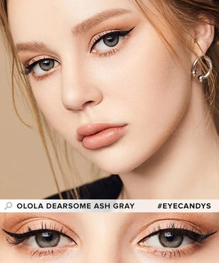 A close-up of a woman wearing Olola Dearsome Ash Grey contact lenses by Olola for dark eyes, with winged eyeliner, nude lipstick, and hoop earrings. Bottom shows a detailed view of her eyes. Text: "Olola Dearsome Ash Grey #Eyecandys.