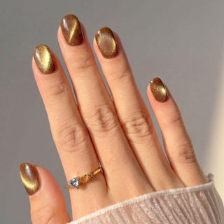 A hand with short, oval-shaped EyeCandys Diet Cola Press-On Nails in a shimmering brown and gold cat-eye effect. The person wears a gold heart gemstone ring on the ring finger and a sheer cream-colored sleeve.