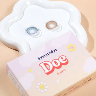 A box labeled "EyeCandys Doe Blue" with floral designs sits before a white tray holding blue and brown natural-iris contact lenses by EyeCandys, all set on a light peach background.