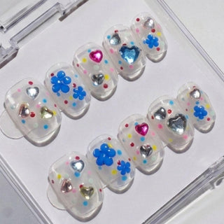 EyeCandys Dot Cake Press-On Nails feature a clear base with colorful polka dots, metallic heart studs, and 3D blue flowers, all neatly packed in a transparent case.