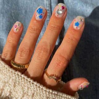 A hand with short nails wears EyeCandys Dot Cake Press-On Nails, featuring colorful gel art—blue flowers, rhinestones, hearts, and multicolored dots. Three gold rings and a cream knit sweater are worn; denim forms the background.