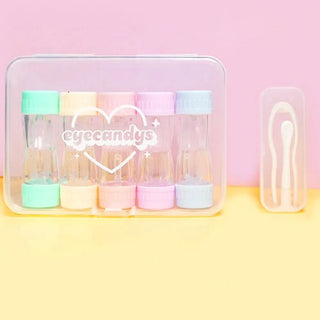 The EyeCandys Cotton Candy Colored Contact Lens Case Travel Kit includes six pastel cases with "eyecandys" text, plus a travel case holding white tweezers and a stick, all set against a vibrant pink and yellow background.