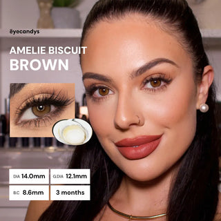 A woman with long dark hair and bold makeup wears EyeCandys Amelie Biscuit Brown contact lenses for a subtle brown eye enhancement. An inset shows a close-up of her eye, product specs, and the text "EYE CANDYS AMELIE BISCUIT BROWN.