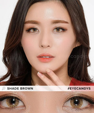 A woman with long dark hair wears natural makeup, including light blush, defined brows, peach lipstick, and highlights. She showcases EyeCandys Shade Brown contact lenses by EyeCandys in a close-up of her brown eyes.