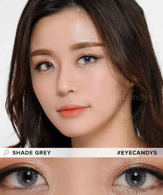 A woman with long dark hair wears EyeCandys Shade Grey cosmetic contact lenses. Her makeup is natural with shimmery eyeshadow and soft coral lips. A close-up shows the contacts on dark eyes. Text: "SHADE GREY #EYECANDYS".