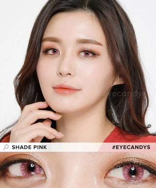 A woman with fair skin and straight dark hair wears EyeCandys Shade Pink contact lenses and subtle makeup. She's in a red top, gently touching her face. A close-up below highlights her striking pinkish-purple eyes.
