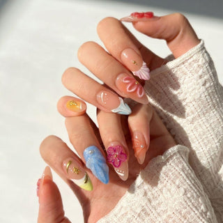 A hand wears EyeCandys Flora Bora Press-On Nails—long, almond-shaped tips with colorful 3D flowers, shells, gold accents, and abstract patterns—held against a light background in a textured white sleeve for a salon look at home.