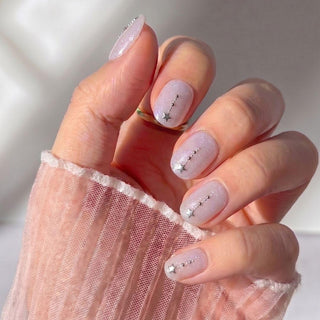 A hand wears EyeCandys Gemini Press-On Nails—light pink, sheer, and glossy with tiny silver star and dot accents. The wrist and fingers are draped in sheer, textured light pink fabric.