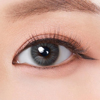 Close-up of a fair-skinned human eye with natural makeup, long lashes, defined brow, and subtle winged eyeliner. The iris looks grayish-blue, enhanced by Olola Heiress Grey contact lens from Olola.