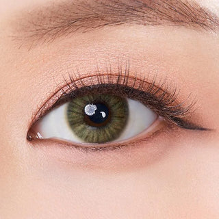 A close-up of a human eye wearing Olola Heiress Olive by Olola shows olive contact lenses, long lashes, and peach eyeshadow. The smooth, well-lit skin highlights how these prescription lenses beautifully enhance dark eyes.