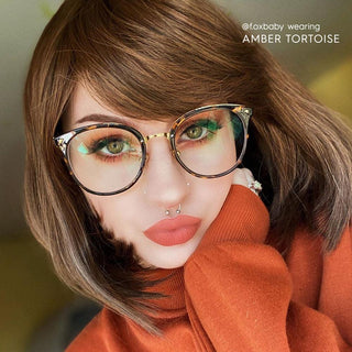 A person with short brown hair, bold makeup, a septum piercing, and an orange sweater poses with their hand on their cheek. Text reads "@foxbaby wearing EyeCandys Infinity Oversized Oval in Amber Tortoise.