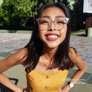 A young woman with straight dark hair and clear cat-eye vintage eyeglasses, wearing a mustard yellow sleeveless top, smiles brightly outdoors. Sunlight highlights her face. Text reads: "KATHLOUISE in EyeCandys Infinity Oversized Oval.