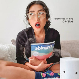 A young woman with a surprised look wears EyeCandys Infinity Oversized Oval vintage eyeglasses, sitting cross-legged on a couch and holding a "Frankie" magazine. She sports bright blue lipstick. Text reads "@kathlouise wearing EyeCandys.
