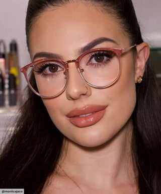 A woman with long dark hair and bold eyebrows wears EyeCandys Infinity Oversized Oval pink glasses and gold stud earrings, looking at the camera with natural makeup; blurred makeup items appear in the background.
